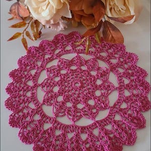 May include: A pink crocheted doily with a delicate floral design. The doily is circular and has a scalloped edge.