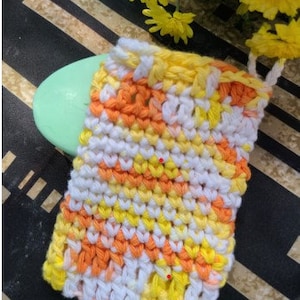 May include: A crocheted soap saver in white, yellow, and orange. The soap saver is made with a textured, woven pattern.