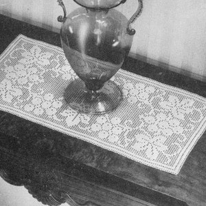 May include: A white crocheted table runner with a floral design. The runner is on a dark wooden table with a clear glass vase.