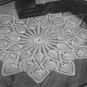 May include: A white crocheted doily with a starburst design. The doily is laid out on a wooden table.