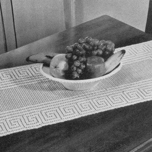 Op de afbeelding: Een witte gehaakte tafelloper met een geometrisch patroon, met in het midden een kom met fruit. De kom bevat druiven, peren en bananen.