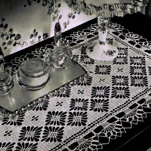May include: A white crocheted table runner with a square pattern and a scalloped edge. The runner is on a dark surface with a glass tray and decorative bottles.