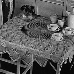 May include: A white crocheted tablecloth with a circular pattern in the center. The tablecloth is laid on a wooden table with a teapot and teacups.