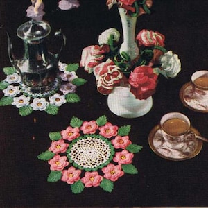 Könnte beinhalten: Drei gehäkelte Untersetzer mit einem floralen Design in Rosa-, Weiß- und Grüntönen. Die Untersetzer liegen auf einer schwarzen Oberfläche mit einer Glaskanne, einer Blumenvase und zwei Teetassen.