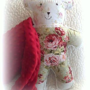 May include: A handmade teddy bear with a floral pattern in red, pink, and green. The bear has a stitched face and is partially covered by a red, textured blanket. The bear is lying on a white, quilted surface.