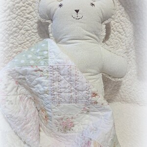 May include: A soft, white teddy bear with embroidered facial features and star accents, nestled under a patchwork quilt. The quilt features floral and polka dot patterns in pastel colors. The bear is on a quilted white surface.