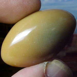 May include: A smooth, polished, oval-shaped stone with a light brown and yellow color. The stone has a subtle sheen and a slightly iridescent quality.