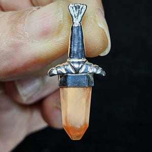 May include: A silver-toned pendant in the shape of a miniature sword. The hilt is detailed with a textured design, and the blade is a translucent orange crystal. The pendant is held against a dark background.