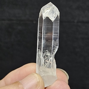 May include: A clear quartz crystal point, held in a hand, against a black background. The crystal is transparent with visible internal structures and a faceted tip. The crystal is approximately 3 inches long.