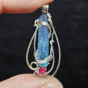 May include: A handcrafted pendant featuring a blue crystal wrapped in silver wire. A small, round, red gemstone is set at the base of the crystal. The pendant is suspended from a silver bail, creating a unique and elegant design.