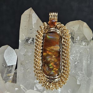 May include: A handcrafted pendant featuring a polished, oval-shaped gemstone with warm brown and iridescent hues. The stone is encased in a gold-colored wire frame with intricate detailing. A small, orange-colored stone is at the top of the pendant.