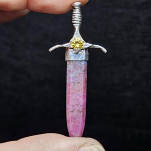 May include: A pendant shaped like a sword, featuring a pink crystal blade and a silver hilt. The hilt has a yellow gemstone at the center. The pendant is held by a hand against a dark background.