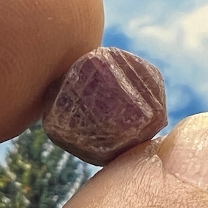 May include: A close-up of a dark purple, faceted gemstone with a rough, natural texture. The gemstone is held between two fingers against a blue sky background.
