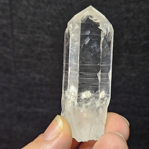 May include: A clear quartz crystal point, showcasing natural facets and a tapered shape. The crystal is held in a hand, revealing its translucent quality and subtle internal textures. The overall appearance is of a raw, unpolished gemstone.