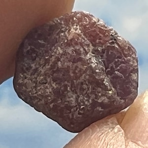 May include: A rough, dark purple gemstone with a textured surface.