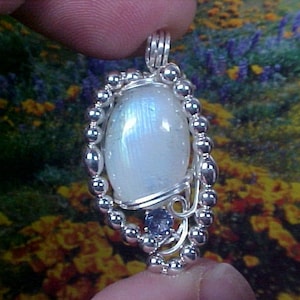 May include: A silver wire wrapped pendant featuring a white moonstone with iridescent blue hues and a smaller blue gemstone accent.