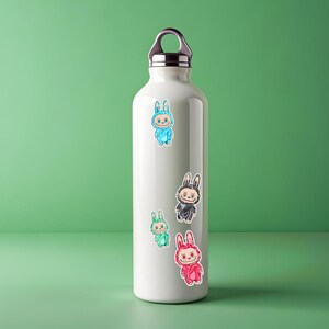 May include: A white insulated water bottle with a silver cap and handle. The bottle is decorated with colorful stickers of cartoon bunnies in blue, black, green, and red outfits. The bottle is on a green surface.