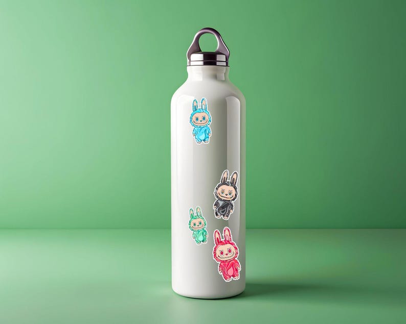 May include: A white insulated water bottle with a silver cap and handle. The bottle is decorated with colorful stickers of cartoon bunnies in blue, black, green, and red outfits. The bottle is on a green surface.