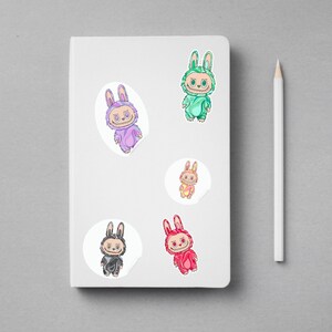 May include: A white notebook with five colorful bunny stickers and a white pencil. The stickers feature cartoon bunnies in various colors, including purple, green, pink, red, and black. The stickers are on a gray surface.