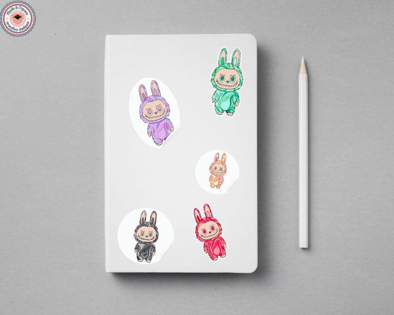 May include: A white notebook with five colorful bunny stickers and a white pencil. The stickers feature cartoon bunnies in various colors, including purple, green, pink, red, and black. The stickers are on a gray surface.