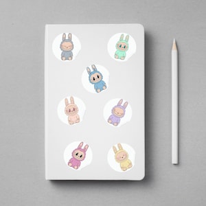 Puede incluir: Un cuaderno blanco con siete pegatinas de conejitos de colores y un l&aacute;piz blanco. Las pegatinas presentan conejitos de dibujos animados en varios colores pastel, incluyendo azul, rosa y amarillo. Las pegatinas est&aacute;n sobre un fondo blanco.