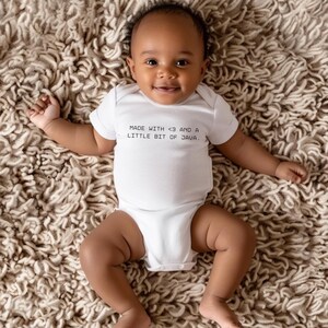 Op de afbeelding: Een lachende baby draagt een witte romper met de tekst "MADE WITH <3 AND A LITTLE BIT OF JAVA." De baby ligt op een getextureerd, lichtbruin oppervlak.