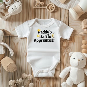 Op de afbeelding: Witte babybody met de tekst "Daddy's Little Apprentice" in zwart, geel en groen. Het ontwerp bevat een bouwvakkershelm, een tandwiel en een boor. De body is omgeven door babyspeelgoed op een houten oppervlak.