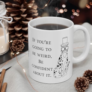 May include: White ceramic mug filled with coffee, displaying a black and white illustration of a cat in a top hat and spectacles. The mug has the text: "If you're going to be weird, be confident about it."