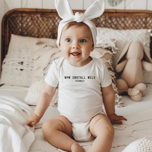 Puede incluir: Un bebé sonriente con un body blanco con el texto "NPM INSTALL MILK <CODE/>" y una diadema de orejas de conejo blancas. El bebé está sentado en una cama con almohadas y un conejito de peluche.
