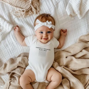 Puede incluir: Un bebé con un body blanco con el texto "CURRENTLY DEBUGGING SLEEP MODE" y una diadema blanca con lazo. El bebé está acostado en una cama con mantas beige y blancas, sonriendo a la cámara.