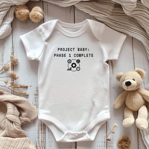 以下が含まれることがあります： 「PROJECT BABY: PHASE 1 COMPLETE」の黒い文字が入った白いベビーボディスーツ。テキストの下には歯車とグラフのグラフィックがあります。背景にはテディベアやその他のベビー用品があります。