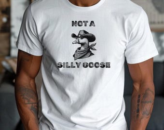 May include: White t-shirt featuring a graphic of a goose wearing a cowboy hat and bandana. The text above the goose reads "NOT A" and below it reads "SILLY GOOSE" in a distressed font. The shirt is a casual wear item.