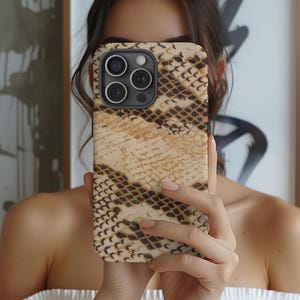 Könnte beinhalten: Eine Smartphone-Hülle mit Schlangenmuster in den Farbtönen Beige, Braun und Schwarz. Die Hülle hat einen Kameraausschnitt mit drei Objektiven und einem Blitz. Die Hülle wird vor dem Gesicht einer Person gehalten.