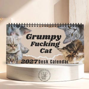 Könnte beinhalten: Ein Tischkalender für 2027 mit schwarzer Spiralbindung, der verschiedene Katzenporträts und den Text Grumpy Fucking Cat 2027 Desk Calendar in Schwarz auf beigem Hintergrund zeigt. Der Kalender enthält ein kreisförmiges Logo mit einem Elefanten.