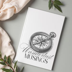May include: A white journal with a detailed black and white compass illustration. The words "Wanderlust Musings" are written in elegant script below the compass. The journal is on a gray surface, with a cream-colored cloth and green leaves in the background.