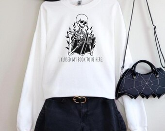 May include: White sweatshirt with a black skeleton graphic reading a book, with the text "I CLOSED MY BOOK TO BE HERE." The sweatshirt is paired with black shorts and a black bat-shaped purse. A black hat hangs above.