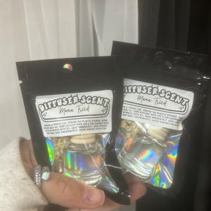 May include: Two black pouches with iridescent foil, each holding a small glass diffuser bottle. The pouches have a white label with the text "DIFFUSER-SCENT Mama Tried". The bottles are clear with wooden tops.