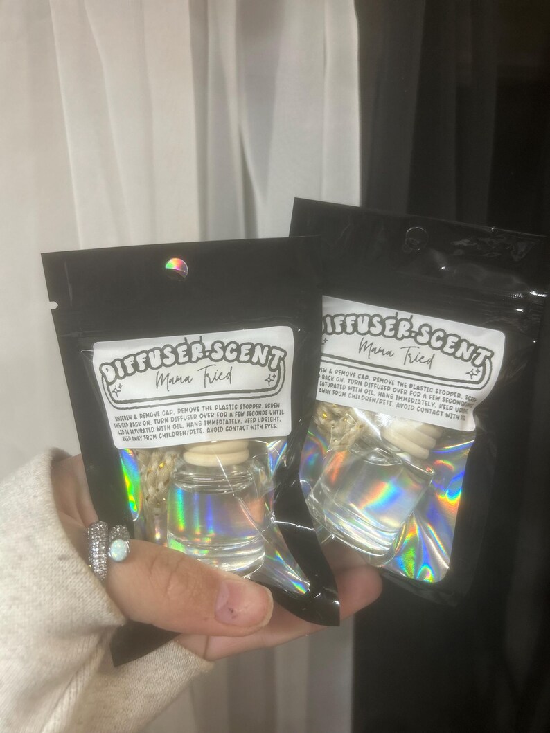 May include: Two black pouches with iridescent foil, each holding a small glass diffuser bottle. The pouches have a white label with the text "DIFFUSER-SCENT Mama Tried". The bottles are clear with wooden tops.
