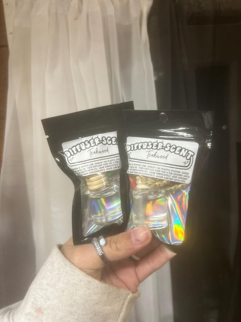 May include: Two Teakwood Diffuser-Scent air fresheners in black packaging. Each has a clear glass bottle with a wooden stopper and a holographic label. The text "DIFFUSER-SCENT Teakwood" is visible on the label.
