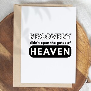 May include: A white greeting card with the text "RECOVERY didn't open the gates of HEAVEN" in black. The card is inside a brown envelope, resting on a wooden tray.