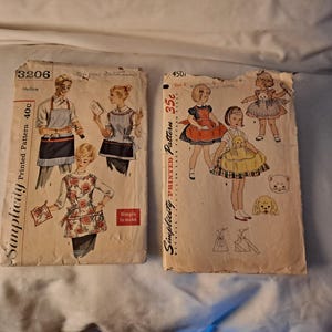 May include: Two vintage Simplicity printed sewing patterns. One pattern features illustrations of aprons, and the other features children's dresses. The patterns are on aged paper with illustrations of people and text.