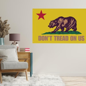 May include: A yellow flag featuring a brown bear walking on green grass, a red star in the upper left corner, and the text "DON'T TREAD ON US" in brown. The flag is displayed on a white wall.