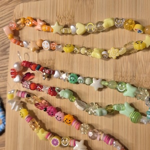 Könnte beinhalten: Vier bunte Perlenarmbänder mit verschiedenen Anhängern, darunter Smiley-Gesichter, Sterne, Herzen und Fruchtformen. Die Armbänder sind auf einer Holzoberfläche angeordnet.