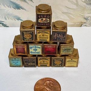 May include: A collection of miniature vintage tea and coffee tins arranged in a pyramid shape. The tins are gold with various labels, including "Tetley", "Twinings", "Flag", "MonoGram", and "Cafe Savoy".