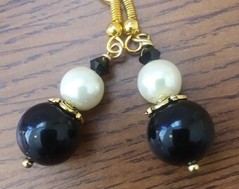 May include: A pair of dangle earrings featuring a black and white bead design. The earrings have gold-colored hooks and accents, with a large black bead at the bottom, a white bead in the middle, and small black accents.