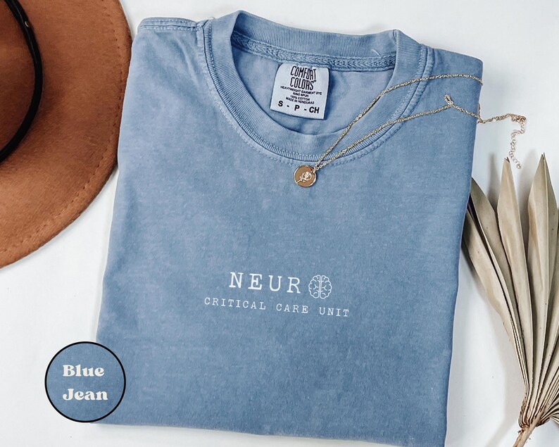 May include: A blue jean Comfort Colors t-shirt with the text "NEUR CRITICAL CARE UNIT" in white. A gold necklace with a pendant is draped across the shirt. A brown hat and dried plant are in the background.