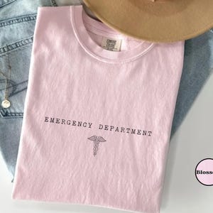 May include: A light pink short-sleeved t-shirt with a crew neckline. The shirt features the black text "EMERGENCY DEPARTMENT" printed above a black caduceus symbol. The inner neck label reads "COMFORT COLORS". A small circular logo with the word "Blossom" is visible.