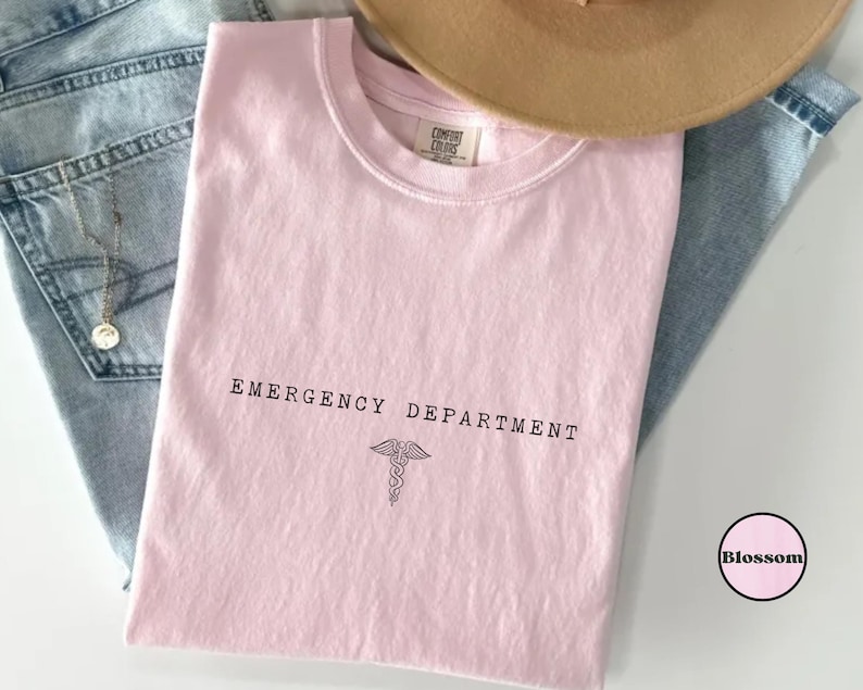 May include: A light pink short-sleeved t-shirt with a crew neckline. The shirt features the black text "EMERGENCY DEPARTMENT" printed above a black caduceus symbol. The inner neck label reads "COMFORT COLORS". A small circular logo with the word "Blossom" is visible.