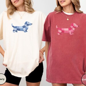 May include: Two t-shirts, one ivory and one crimson, each featuring a dachshund dog design. The dog on the ivory shirt is blue, and the dog on the crimson shirt is pink. The shirts are short-sleeved and have a relaxed fit.