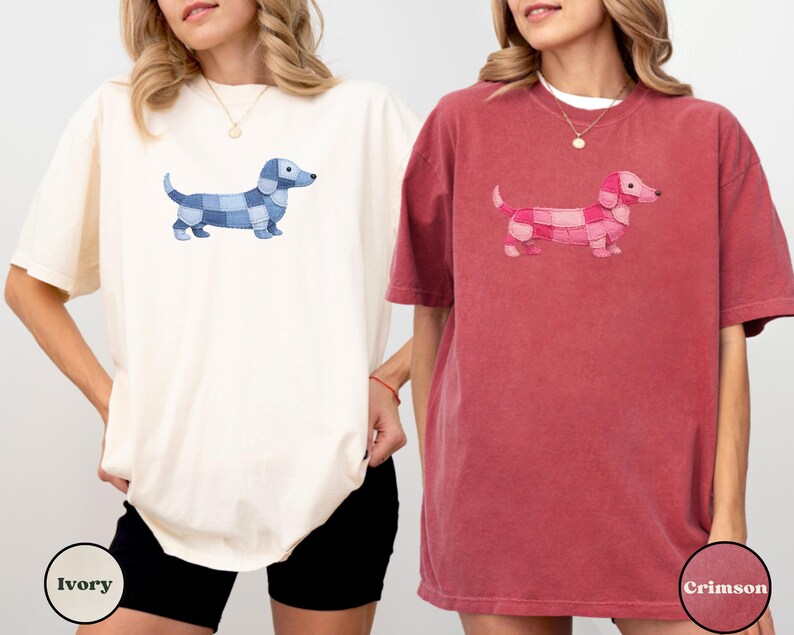May include: Two t-shirts, one ivory and one crimson, each featuring a dachshund dog design. The dog on the ivory shirt is blue, and the dog on the crimson shirt is pink. The shirts are short-sleeved and have a relaxed fit.